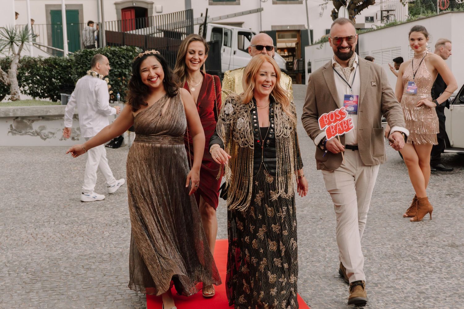 Elegantly dressed group walking a red carpet outdoors at a festive evening event in a scenic location.