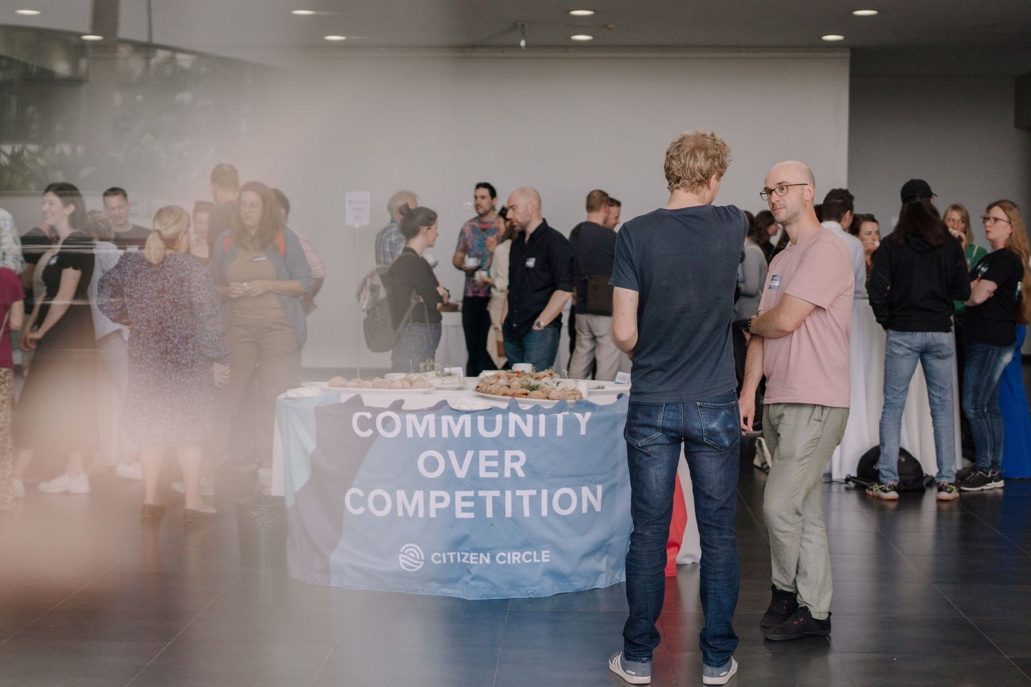 Networking event with a 'Community Over Competition' table banner, attendees mingling in a modern venue.