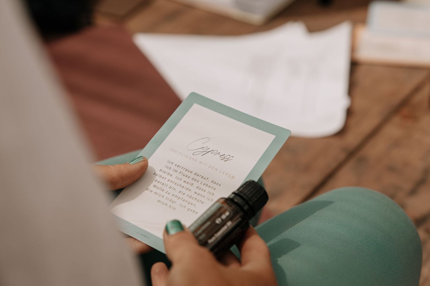 Hands holding a teal card and essential oil bottle on a wooden surface.