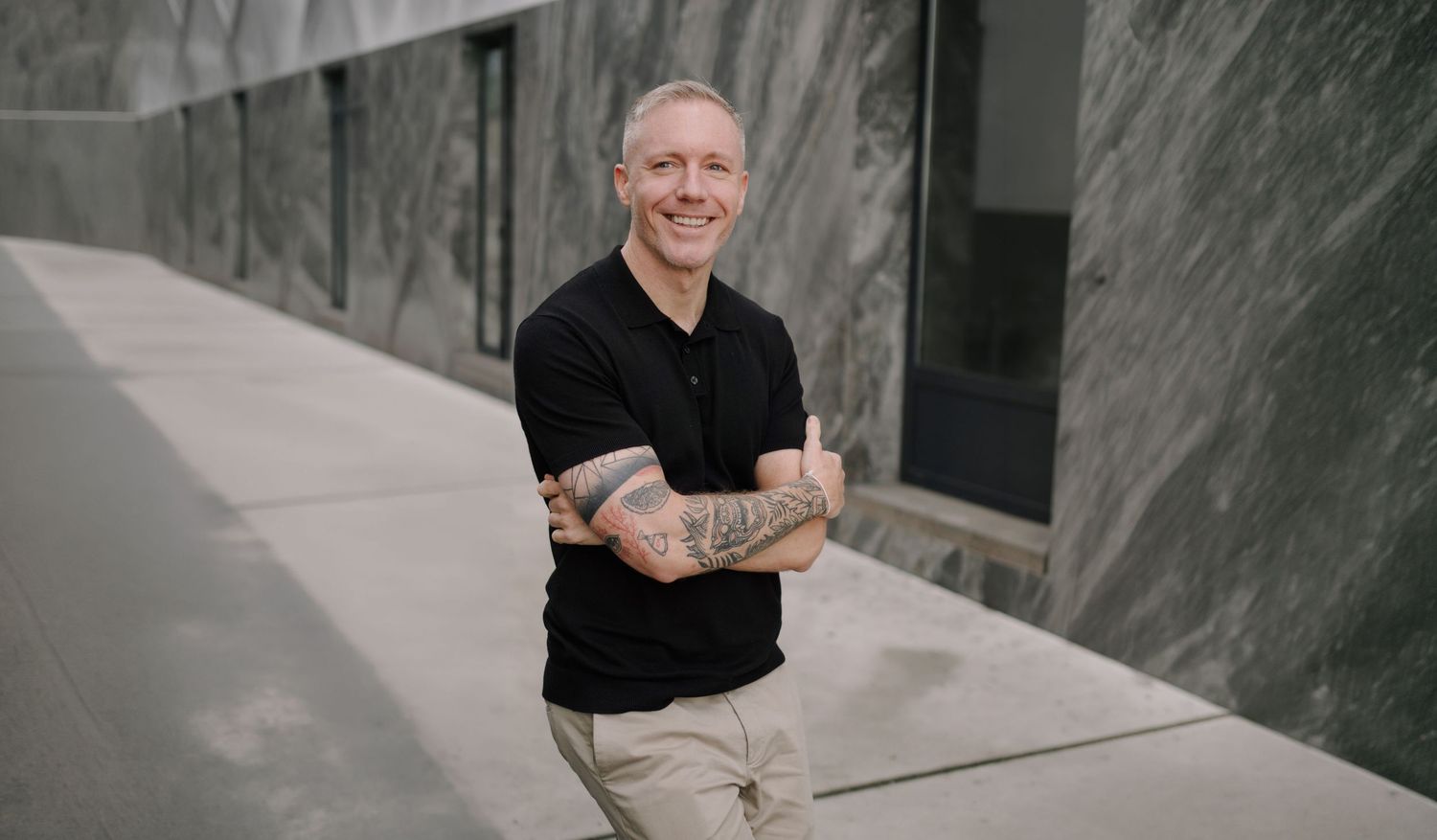 Smiling bald man with tattoos in black polo shirt, arms crossed, standing against modern concrete wall.