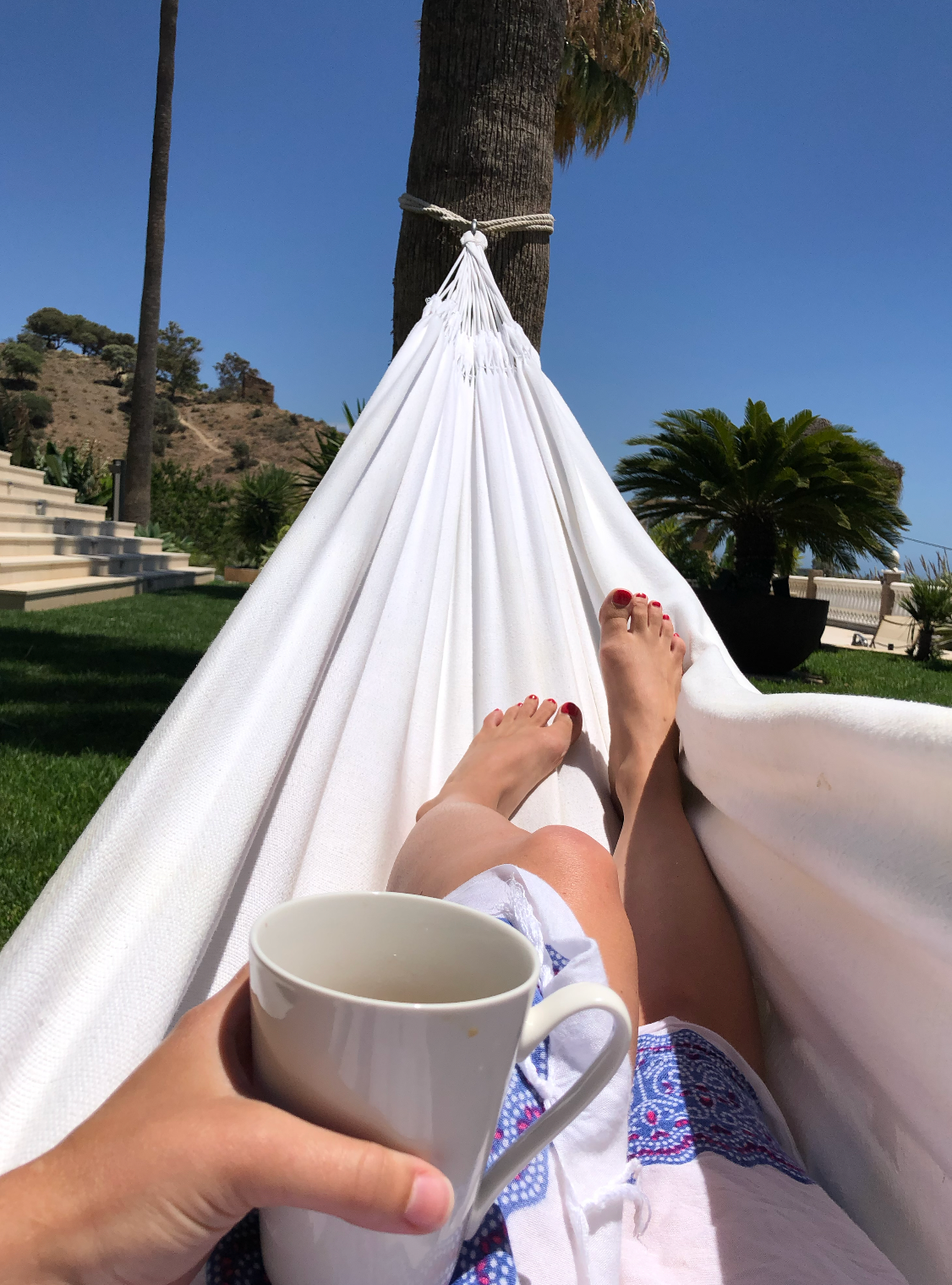 Person relaxing in a white hammock outdoors, holding a coffee mug, with feet up and palm trees in sunny background.
