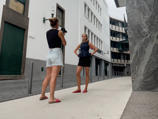 Two women on a city street, one photographing the other posing in a blue dress against a modern building.