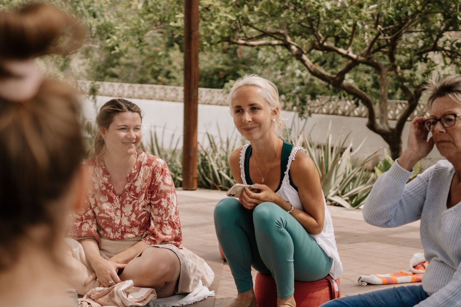 Women sitting outdoors in a casual group conversation, surrounded by lush greenery in a relaxed outdoor setting.