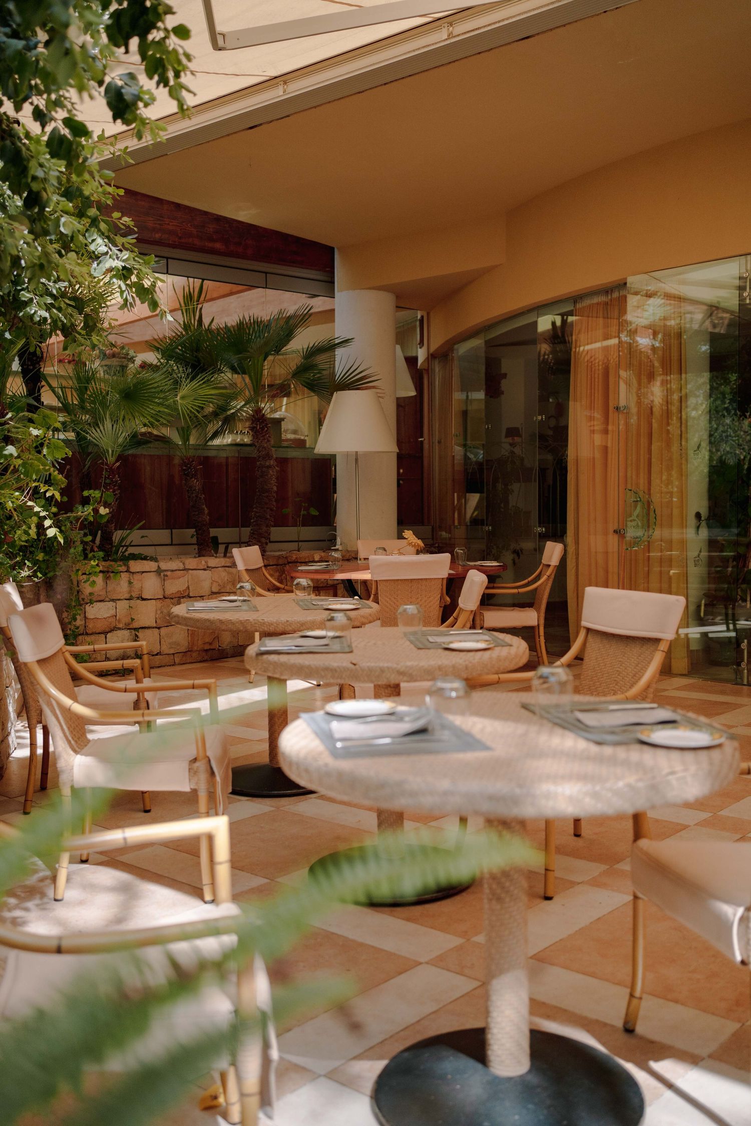 Elegant outdoor patio restaurant with set tables, wicker chairs, checkered tile flooring, and lush palm plants beside a warm golden facade.