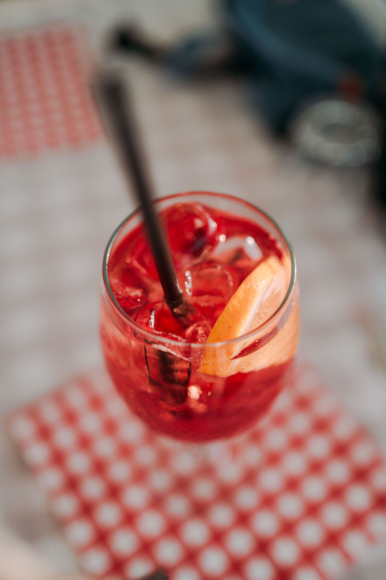 A vibrant red cocktail with ice, orange slice, and straw in a glass on a red checkered napkin.