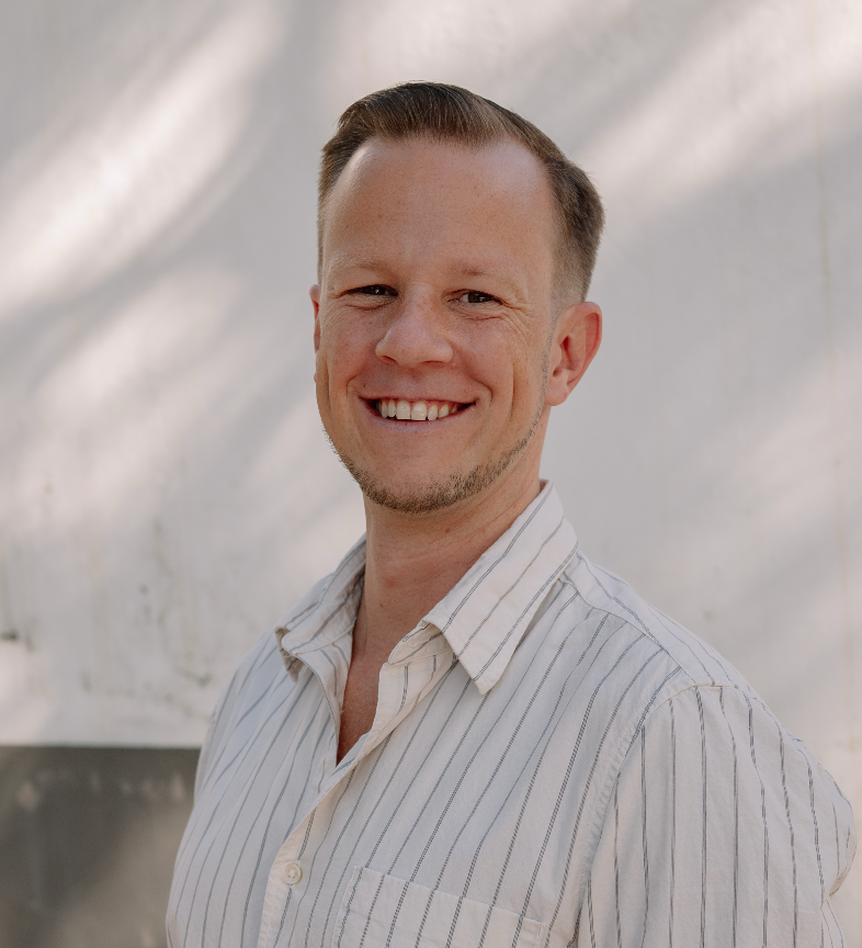 Smiling man with short hair wearing a striped white shirt, standing outdoors in natural light.
