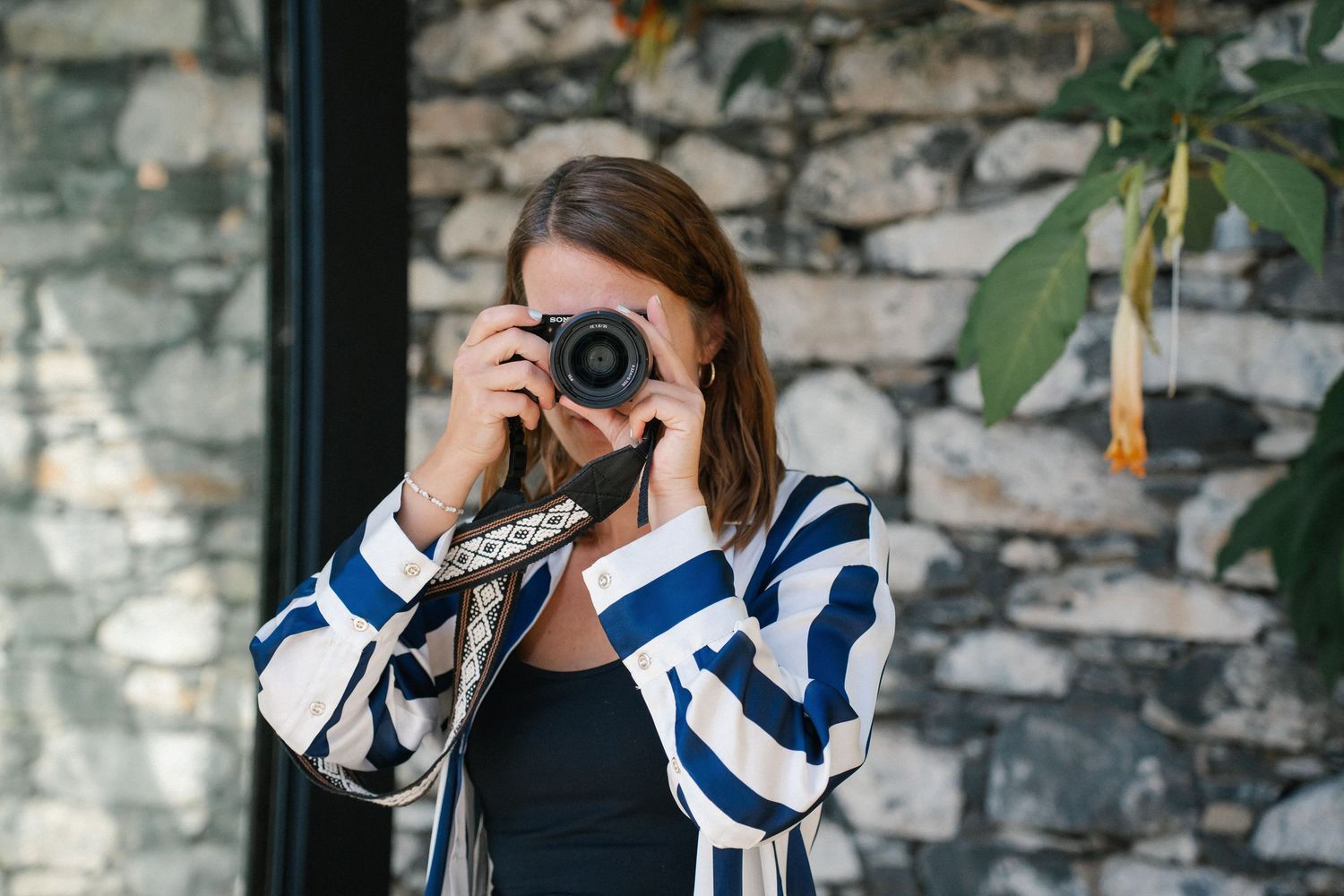 Woman in striped shirt photographing with DSLR camera against rustic stone wall background.