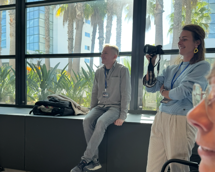 Two people in a modern office lobby with palm trees, one holding a camera, wearing lanyards at a professional event.