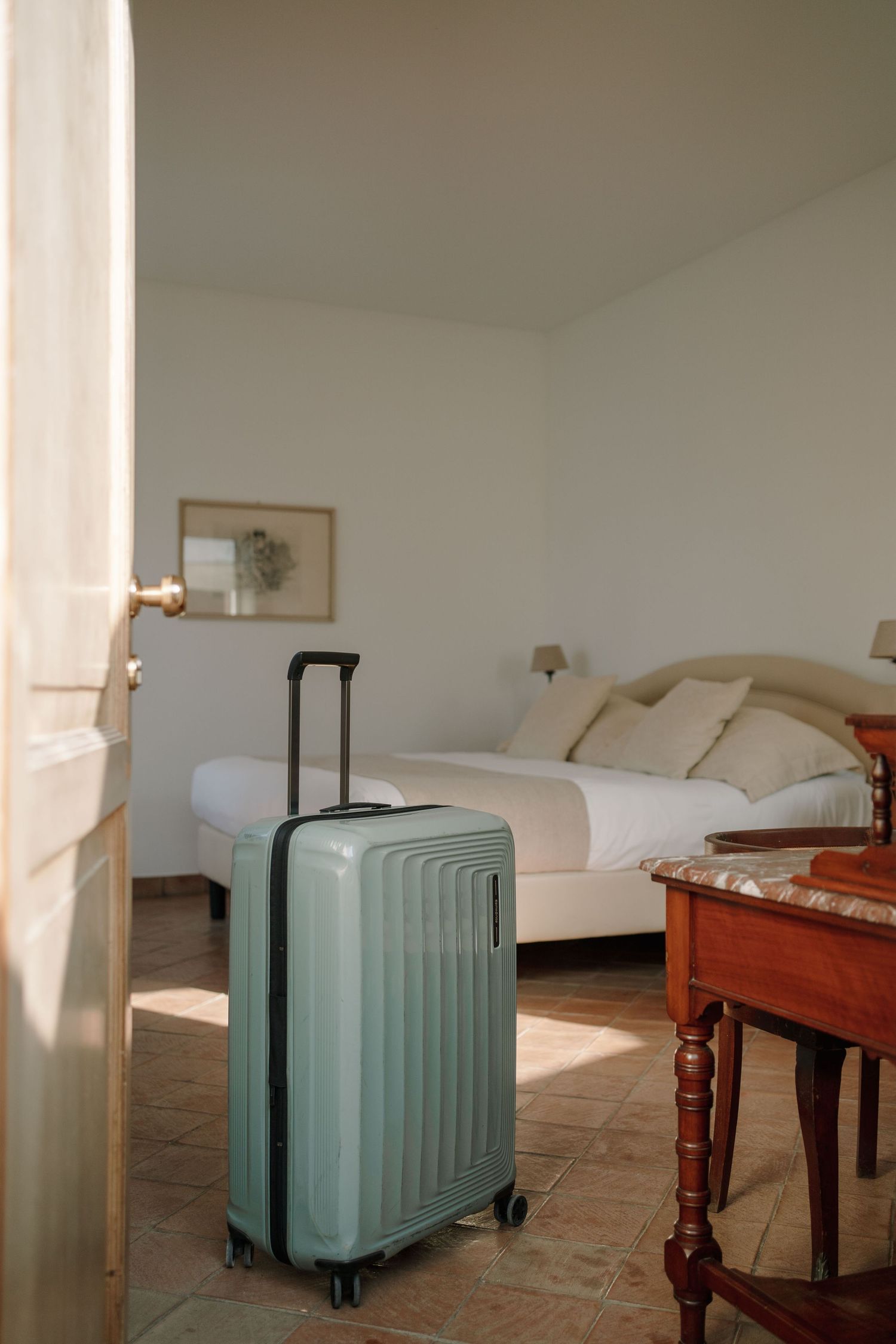 Sage green hardshell suitcase standing in a bright hotel room with a white double bed, terracotta tile floor and wooden table.