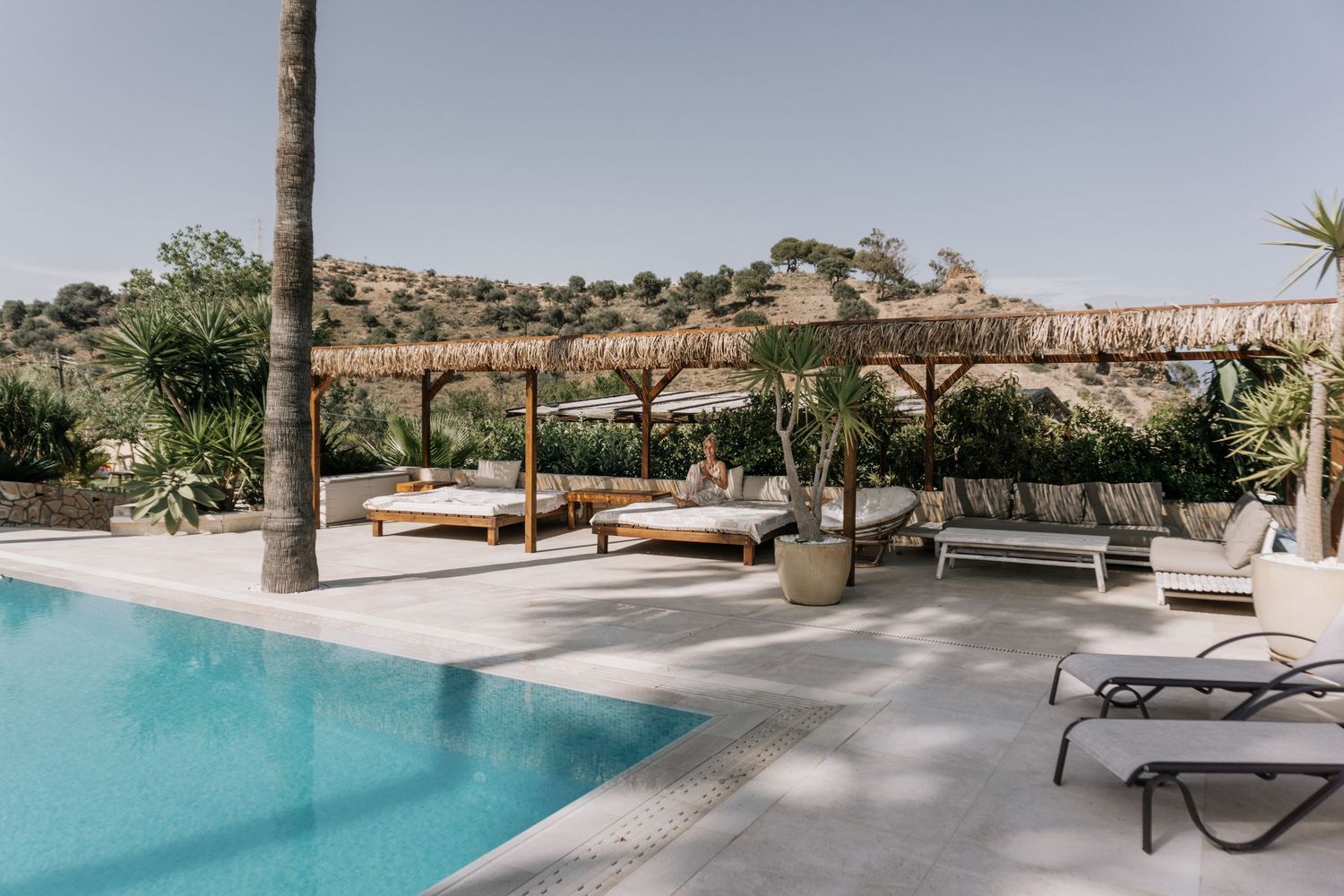 Luxury outdoor pool area with thatched pergola, sun loungers, and lush Mediterranean landscaping under a clear sky.