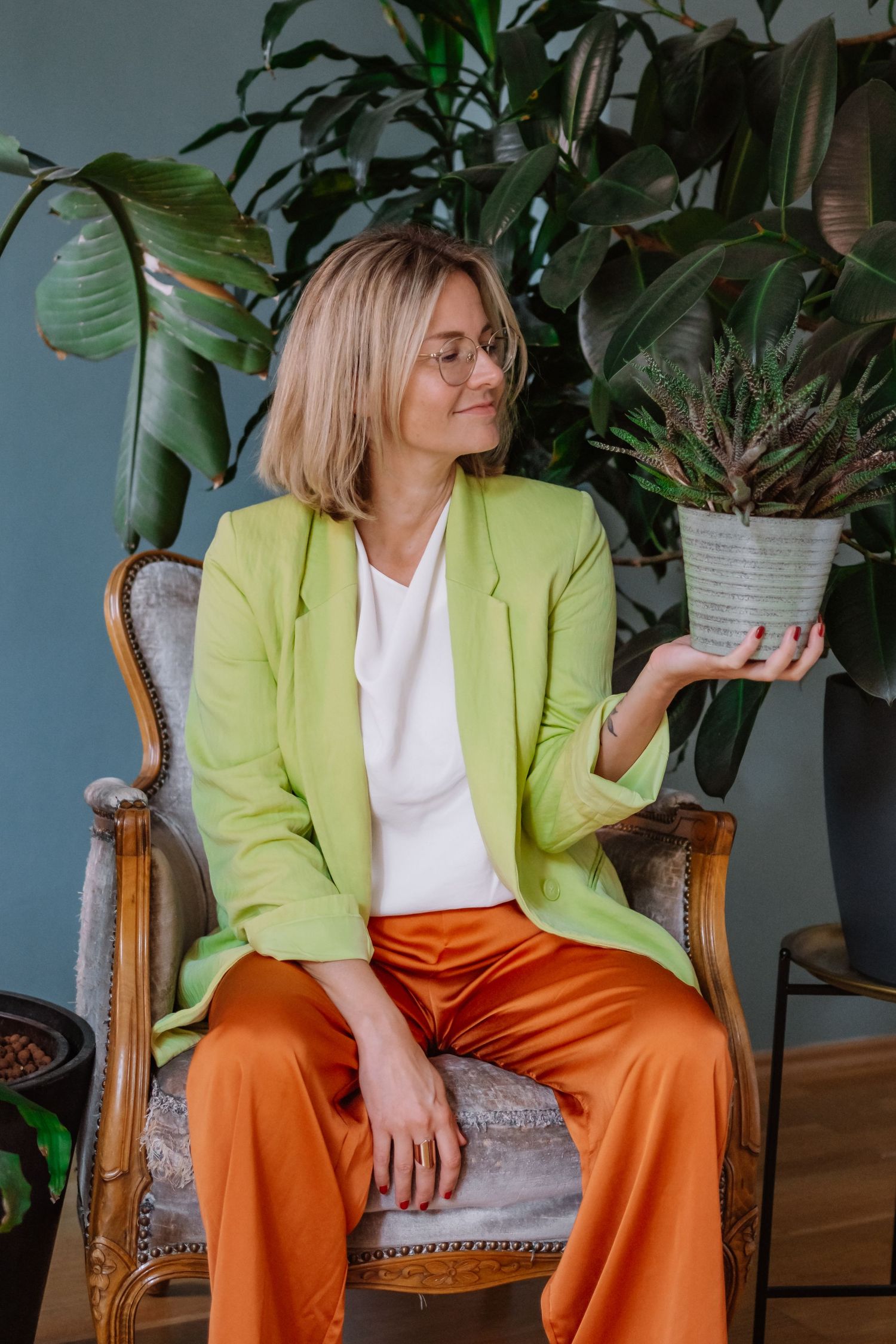 Woman in green blazer and orange pants sits in vintage chair holding a potted plant, surrounded by lush indoor greenery.