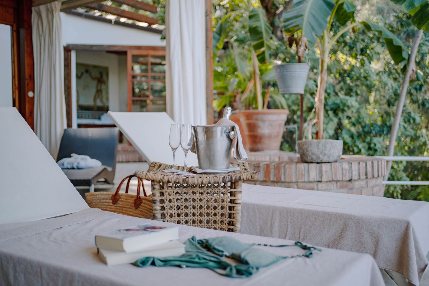 Outdoor terrace lounge with white sun loungers, a wicker basket holding a champagne bucket and glasses, books, and lush tropical plants.
