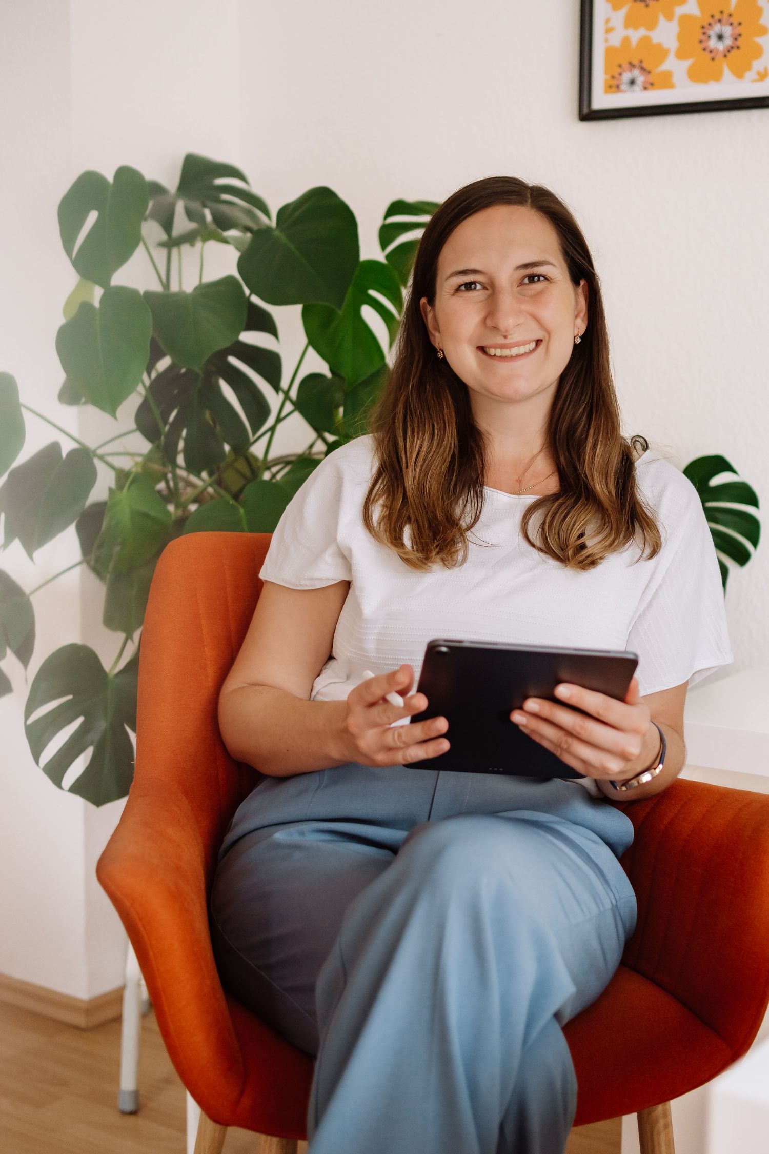 Smiling woman in white top and blue pants sitting in orange chair holding tablet with monstera plant background.