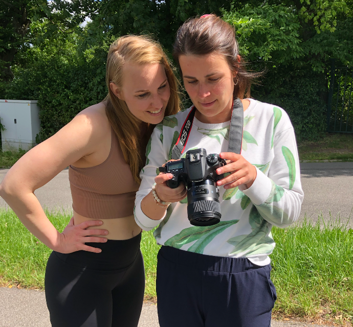 Two young women outdoors reviewing photos on a DSLR camera, one wearing a tan crop top and leggings.