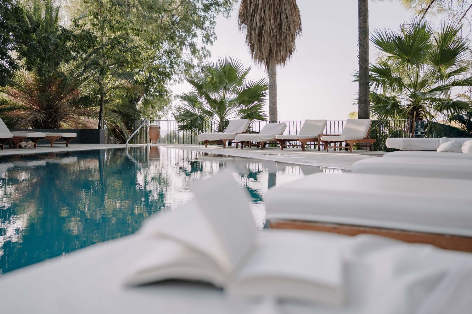 Open book resting on a poolside ledge overlooking a turquoise swimming pool, with lounge chairs and palm trees in the background.