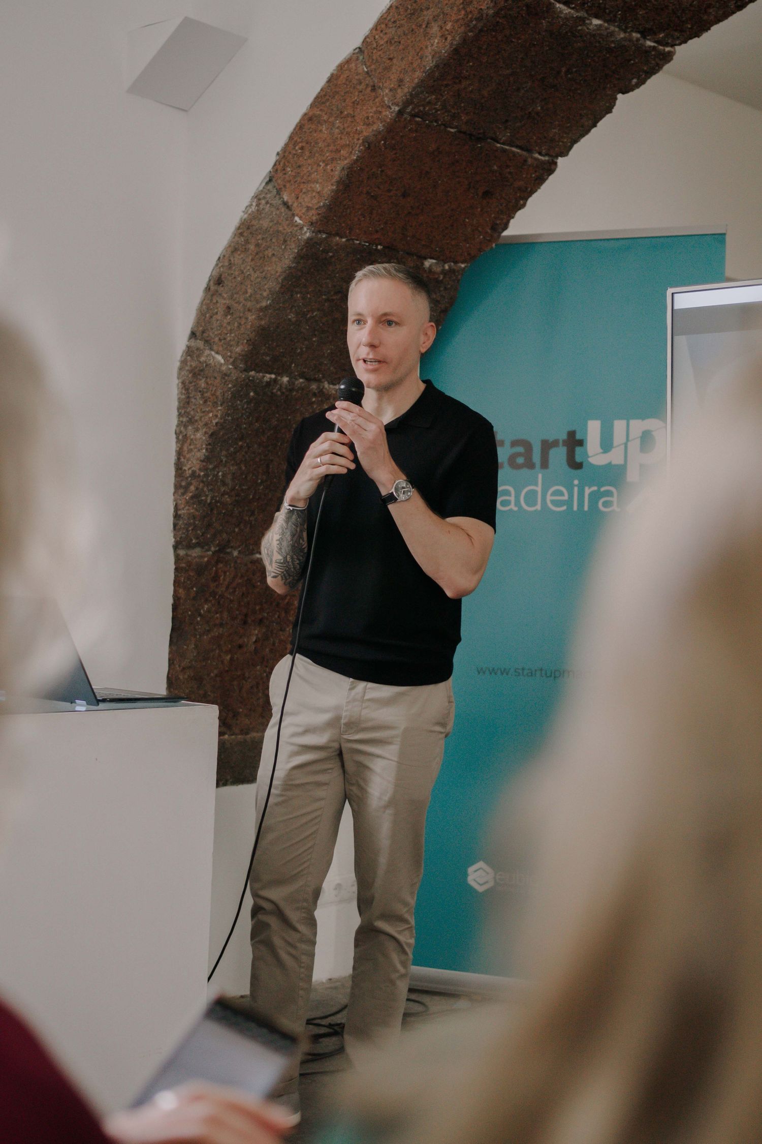 A man in a black shirt speaks into a microphone at a Startup Madeira event under a stone arch.