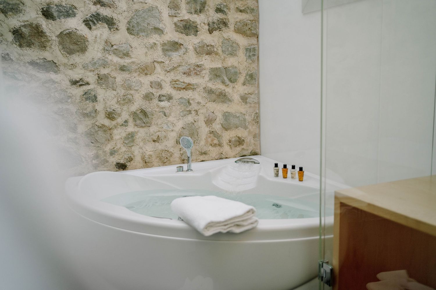Corner whirlpool bathtub with a folded white towel and toiletries beside a rustic natural stone wall in a bright, modern spa-style bathroom.