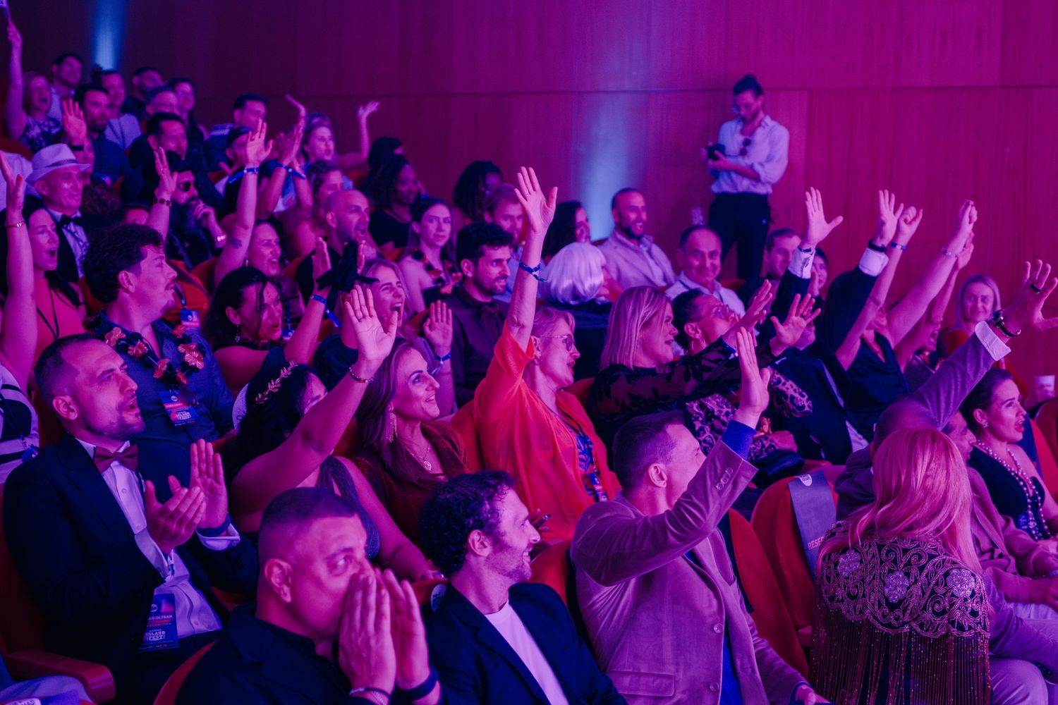 Excited crowd raising hands at a live event under purple stage lighting in a modern auditorium.