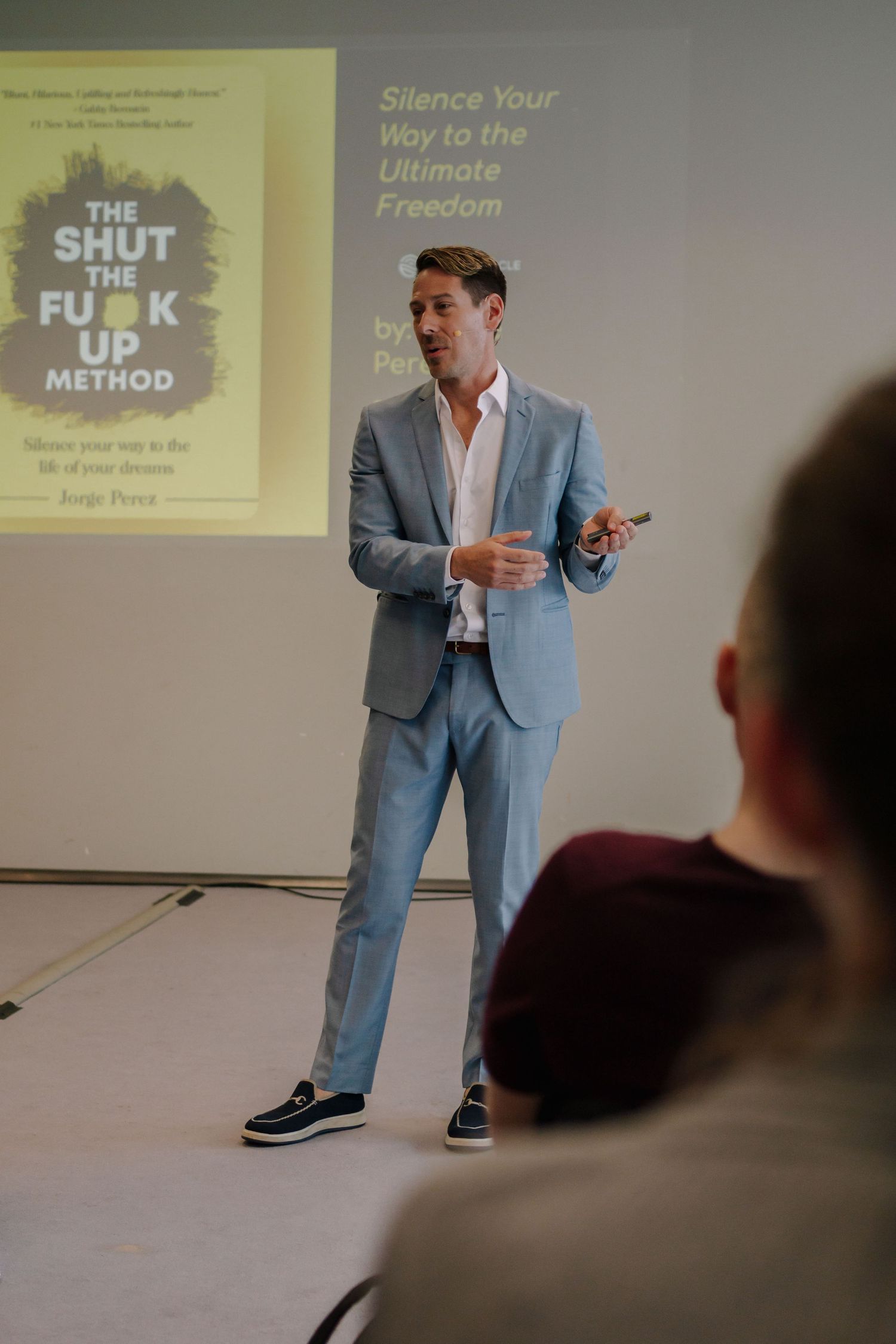 Man in a light blue suit presenting a book called The Shut The Fuck Up Method at a seminar.