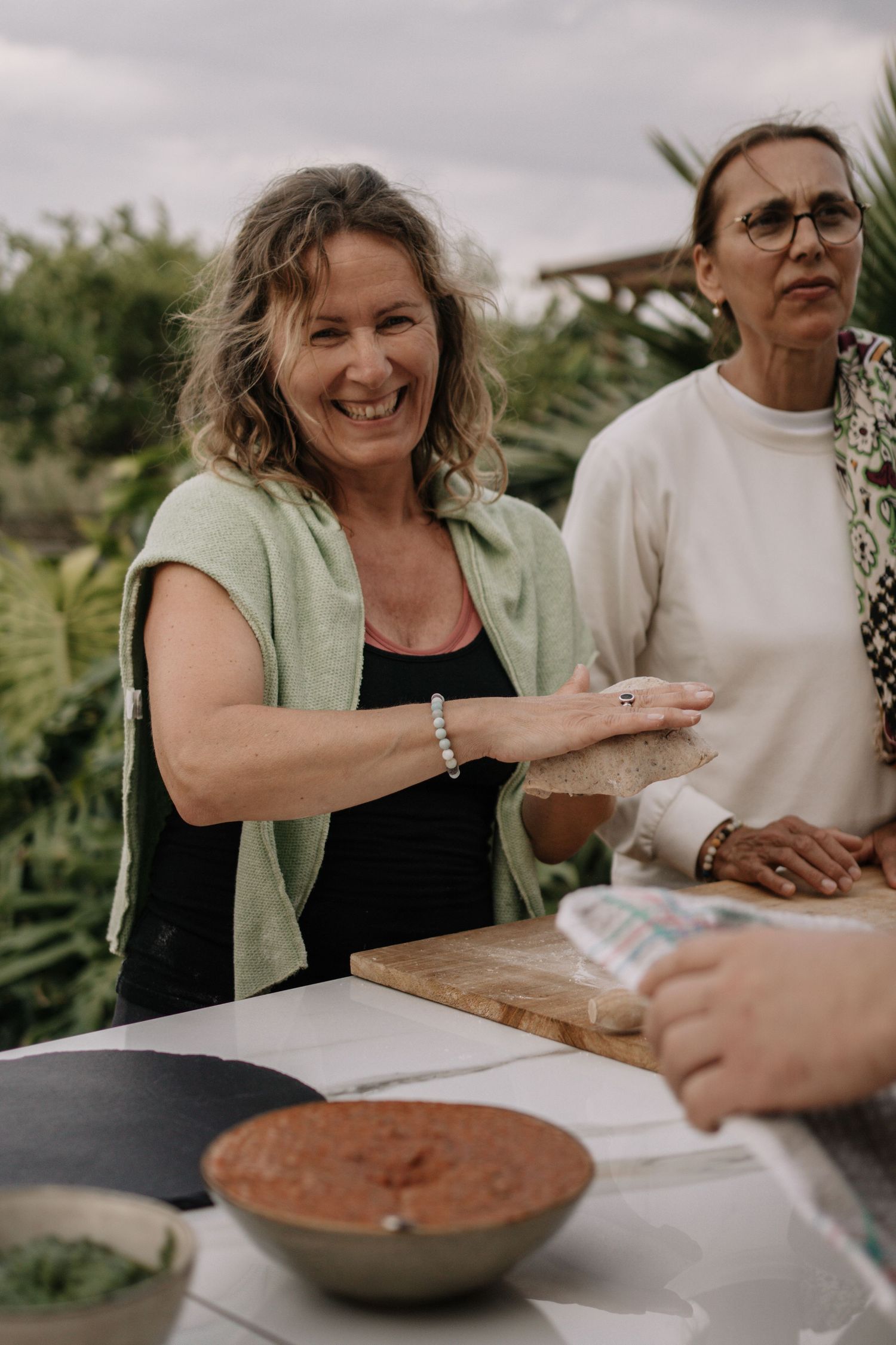 Two women preparing food outdoors, smiling and working with ingredients on a wooden cutting board.