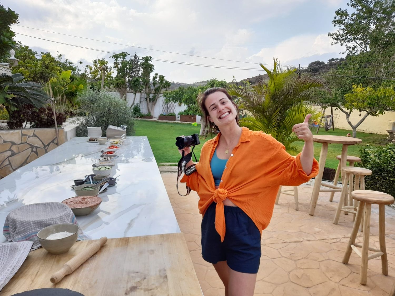 Woman in orange shirt gives thumbs up at outdoor food photography setup with marble table in sunny garden.