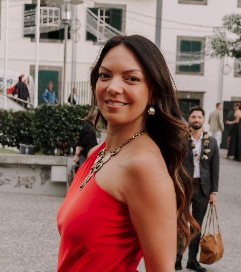 Woman in red dress with gold necklace smiling over shoulder at outdoor event.