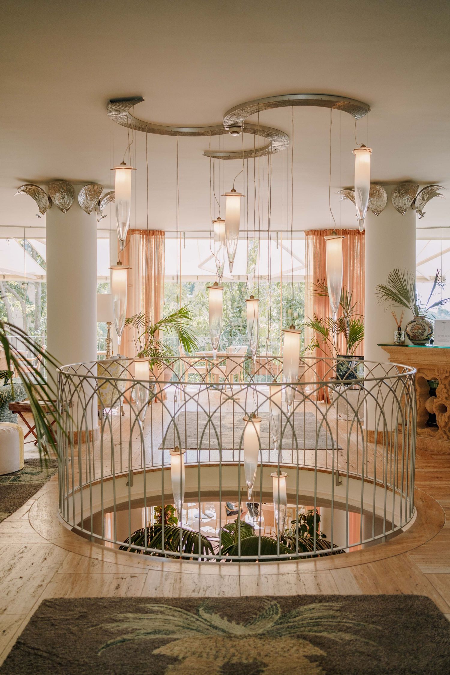 Elegant hotel lobby with a curved wrought-iron railing, hanging glass pendant lights, ornate columns, peach curtains, and tropical plants.