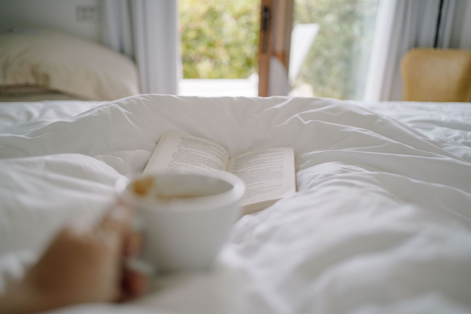 Hand holding a white mug over a rumpled white bed with an open book resting on the duvet beside a sunlit window.