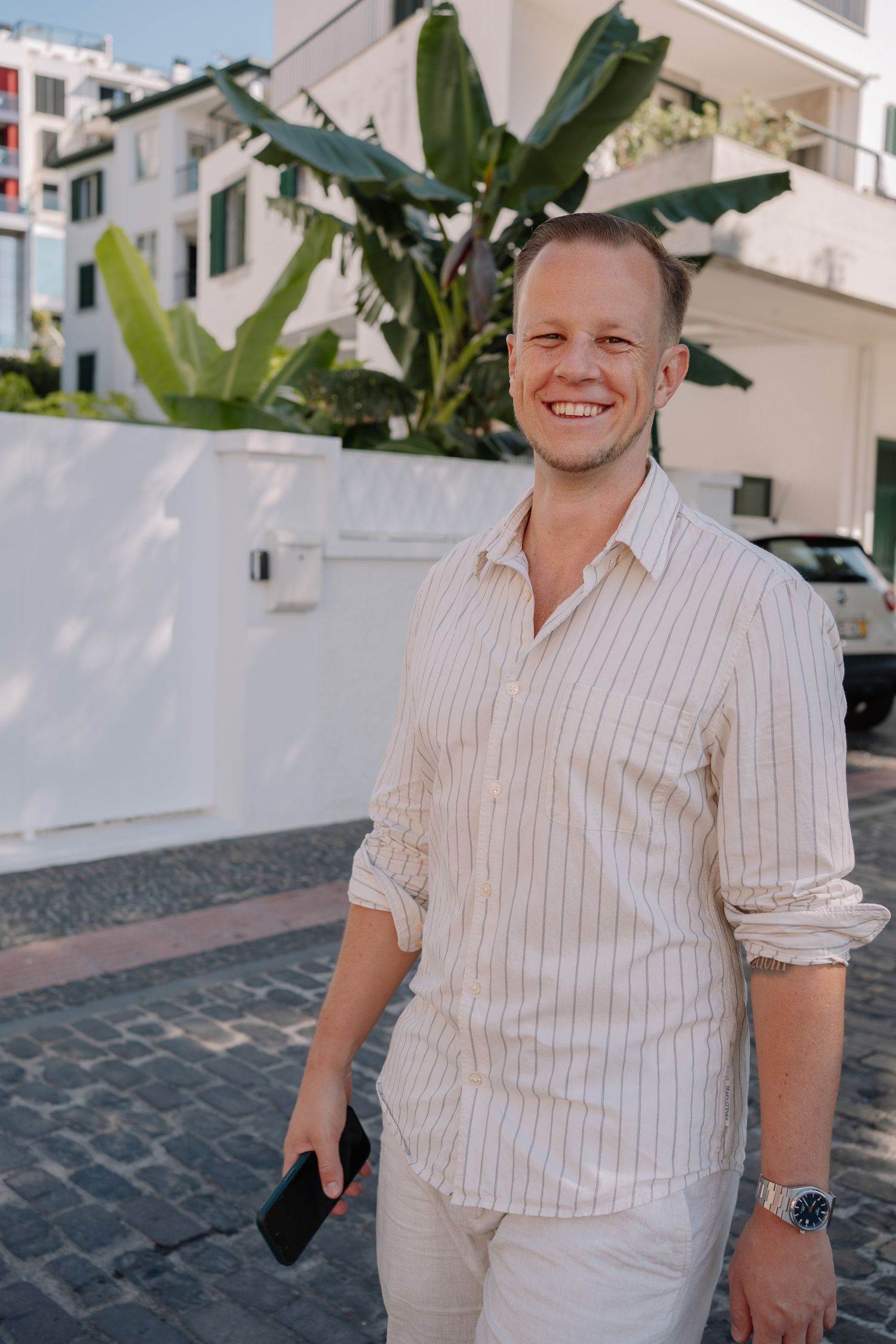 Smiling man in white striped shirt holding phone, standing on cobblestone street with white buildings and tropical plants.