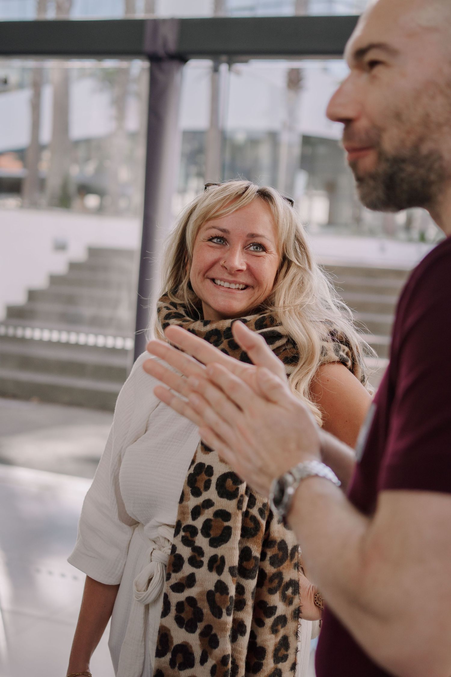 Smiling blonde woman wearing leopard print scarf and white jacket chatting with a man in a maroon shirt outdoors.