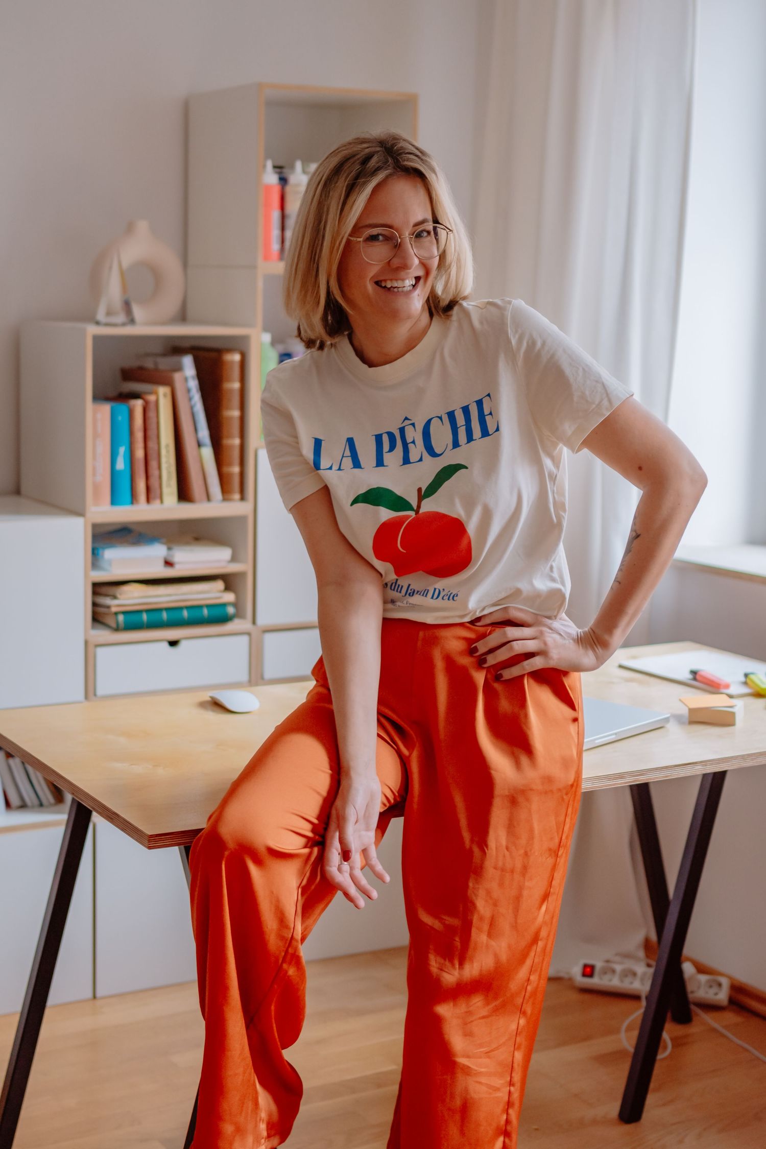 Smiling blonde woman in glasses wearing a white La Peche graphic tee and orange satin trousers, leaning on a desk.