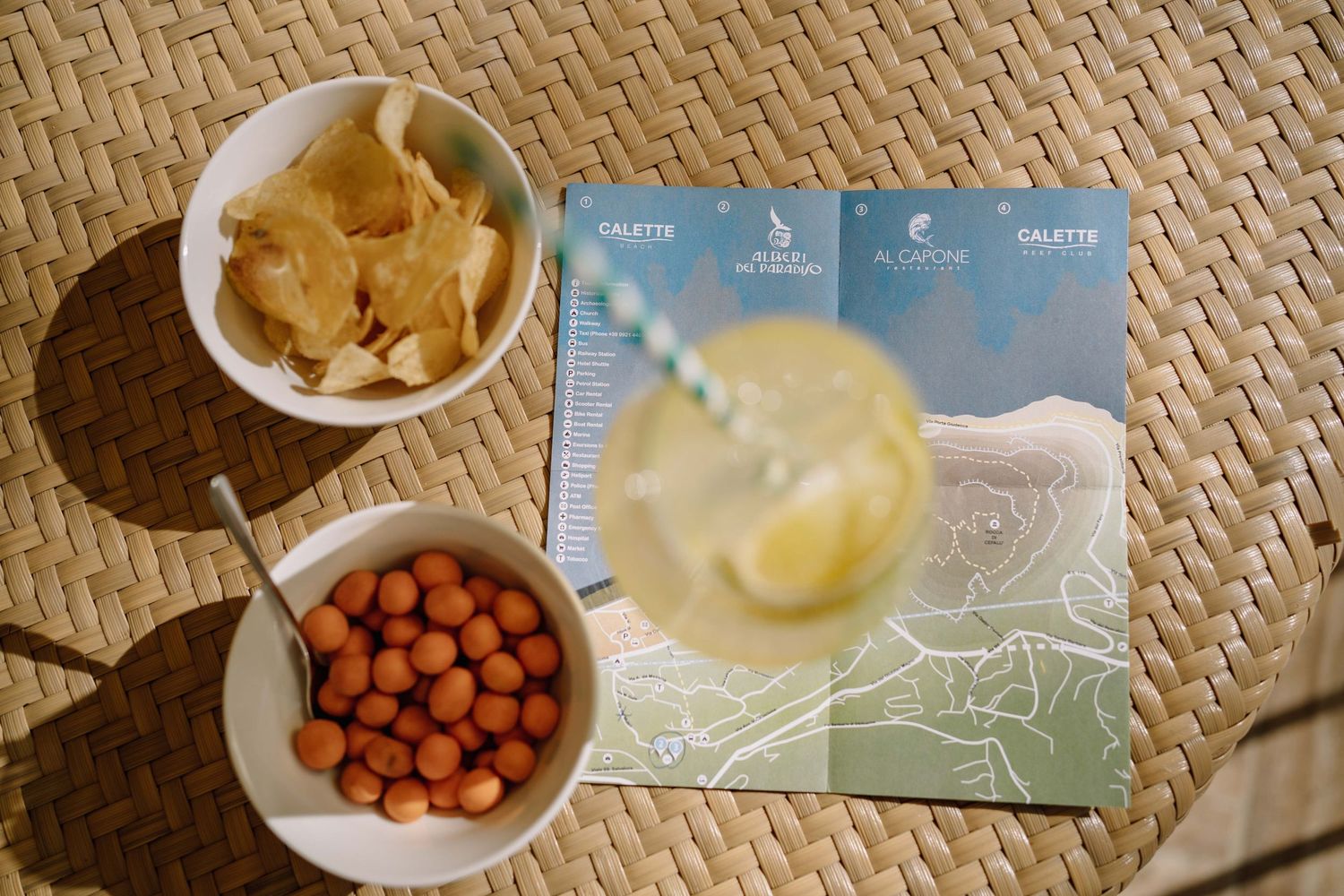 Overhead view of a lemon drink with a striped straw beside bowls of chips and glazed peanuts on a woven table with a menu.