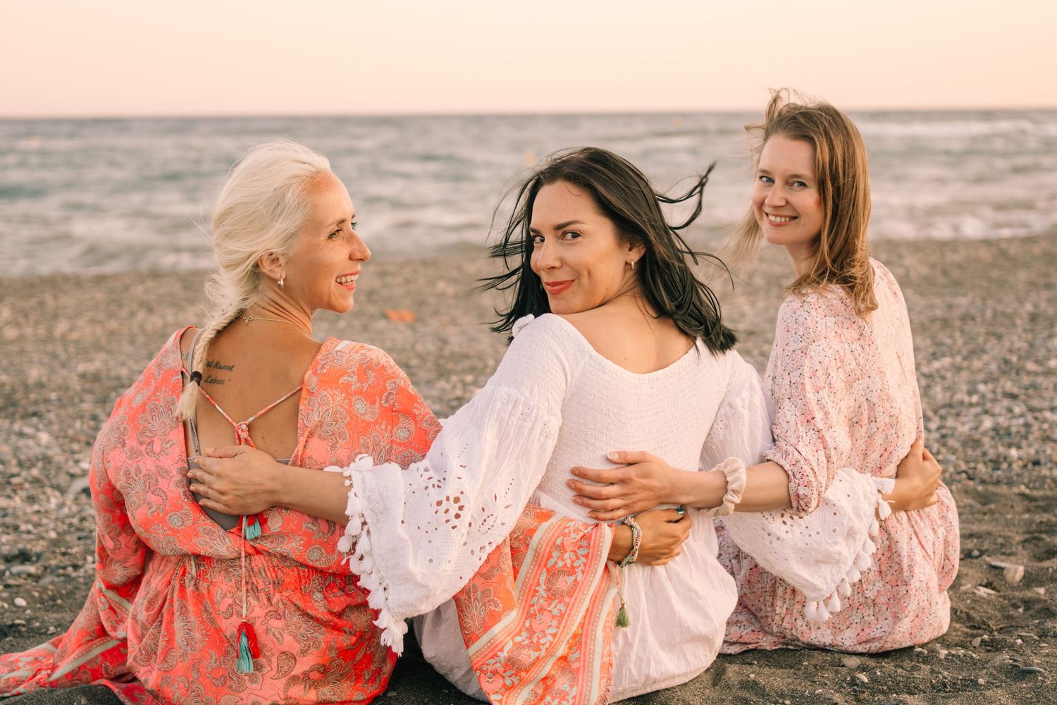 Three women in bohemian dresses sitting together smiling on beach at sunset.