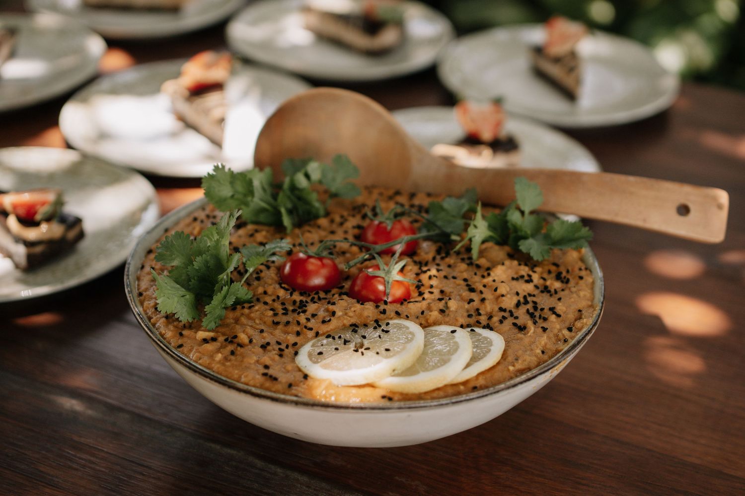 A decorative bowl of hummus topped with cherry tomatoes, black sesame seeds, fresh herbs, and lemon slices on a wooden table.