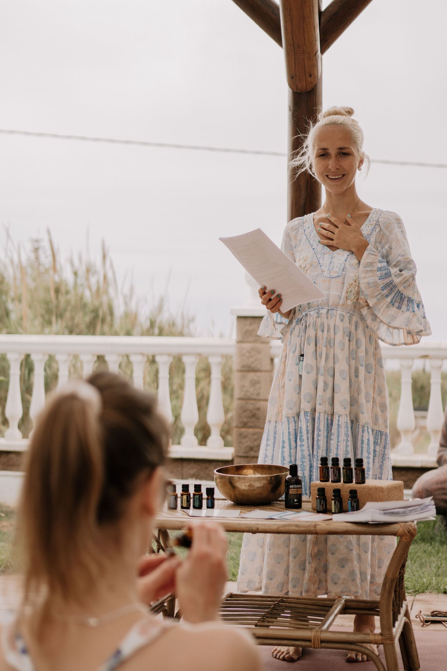 Smiling blonde woman in floral dress leads outdoor wellness workshop with essential oils on terrace.