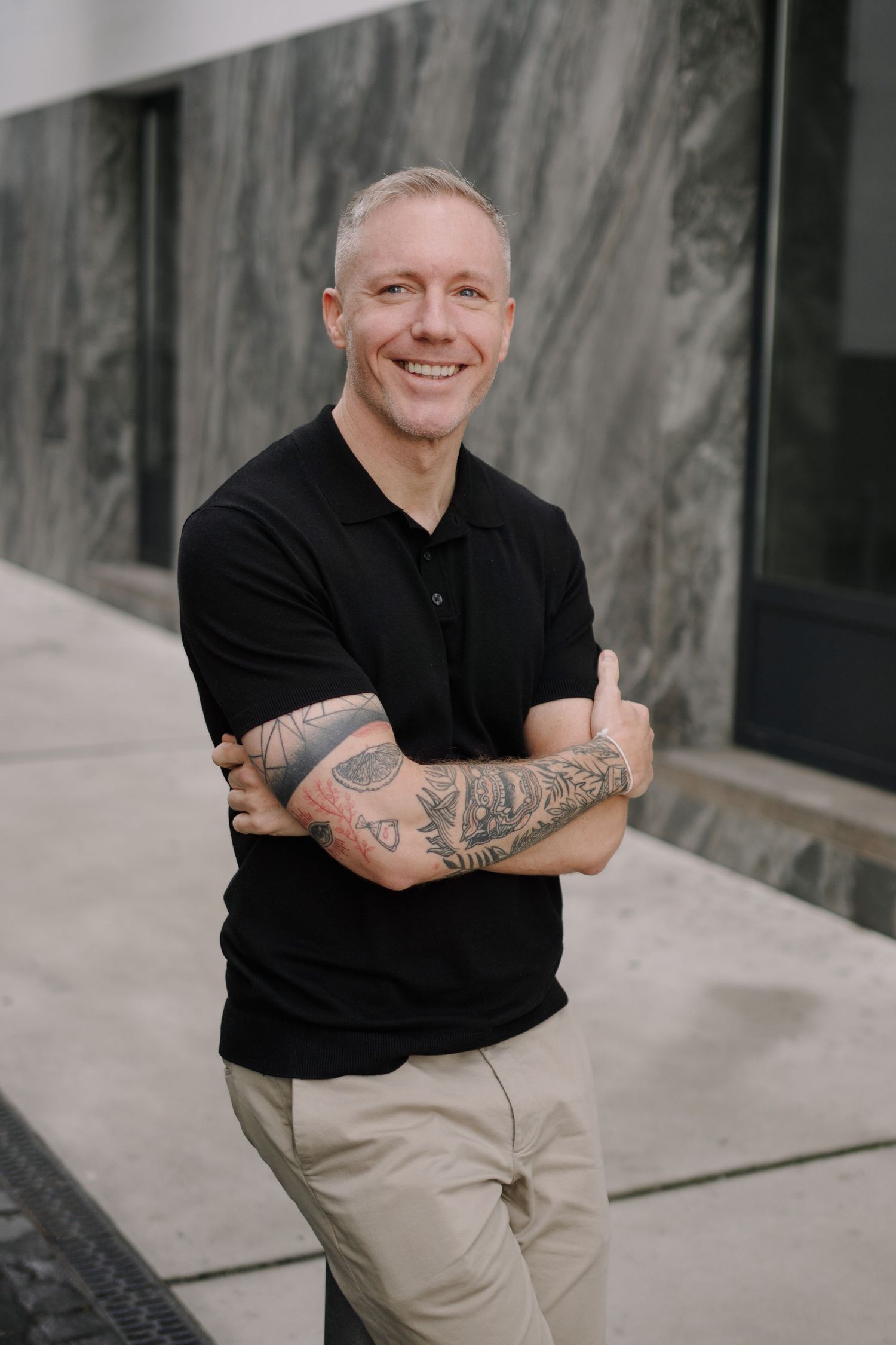 Smiling man with tattoos and short hair wearing a black polo shirt and khaki pants, arms crossed outdoors.