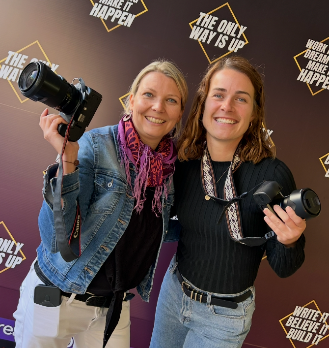 Two women smiling and holding cameras in front of a motivational quote backdrop at a photography event.