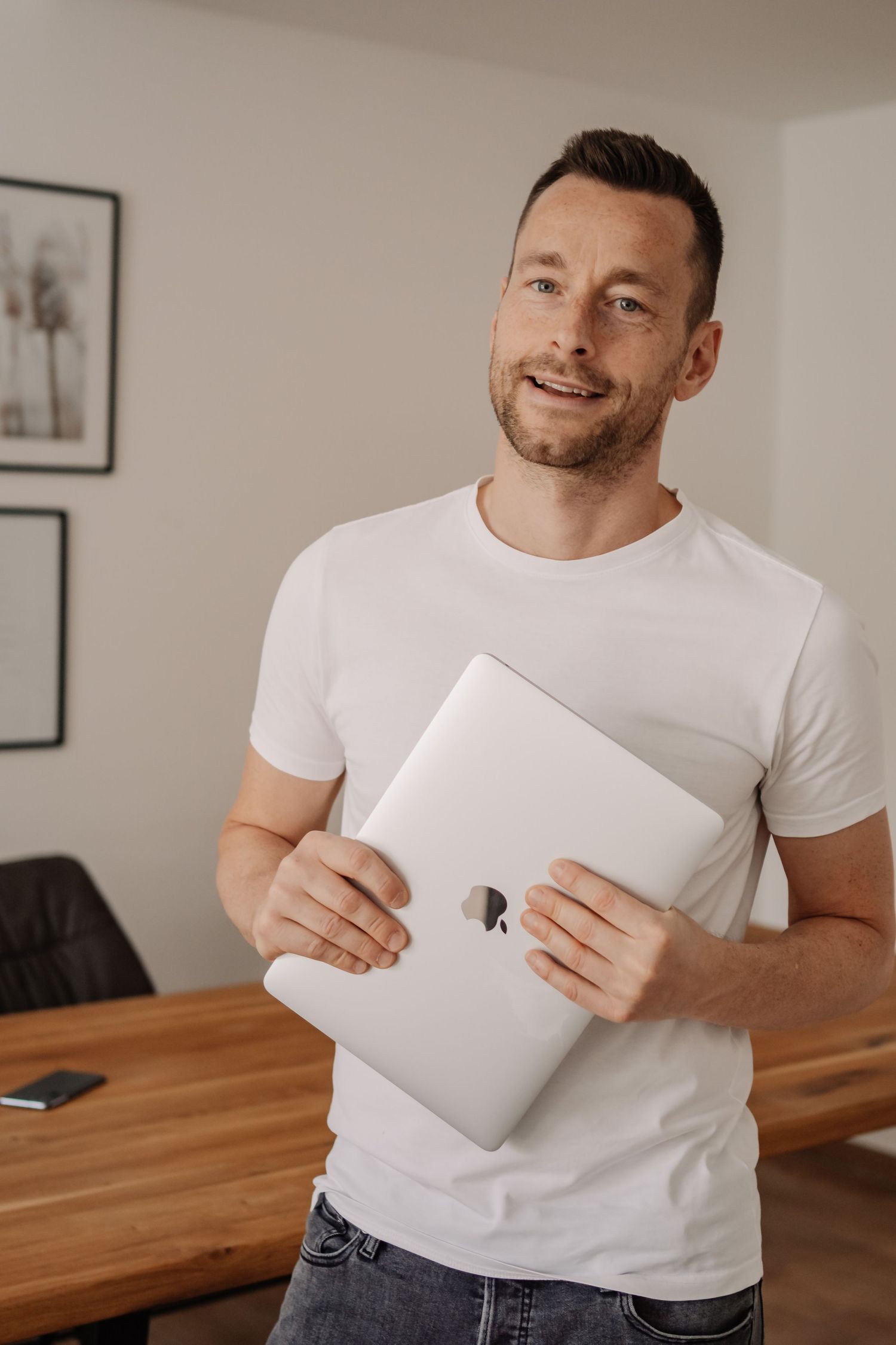 Smiling man in white t-shirt holding a closed Apple MacBook laptop in a modern office setting.