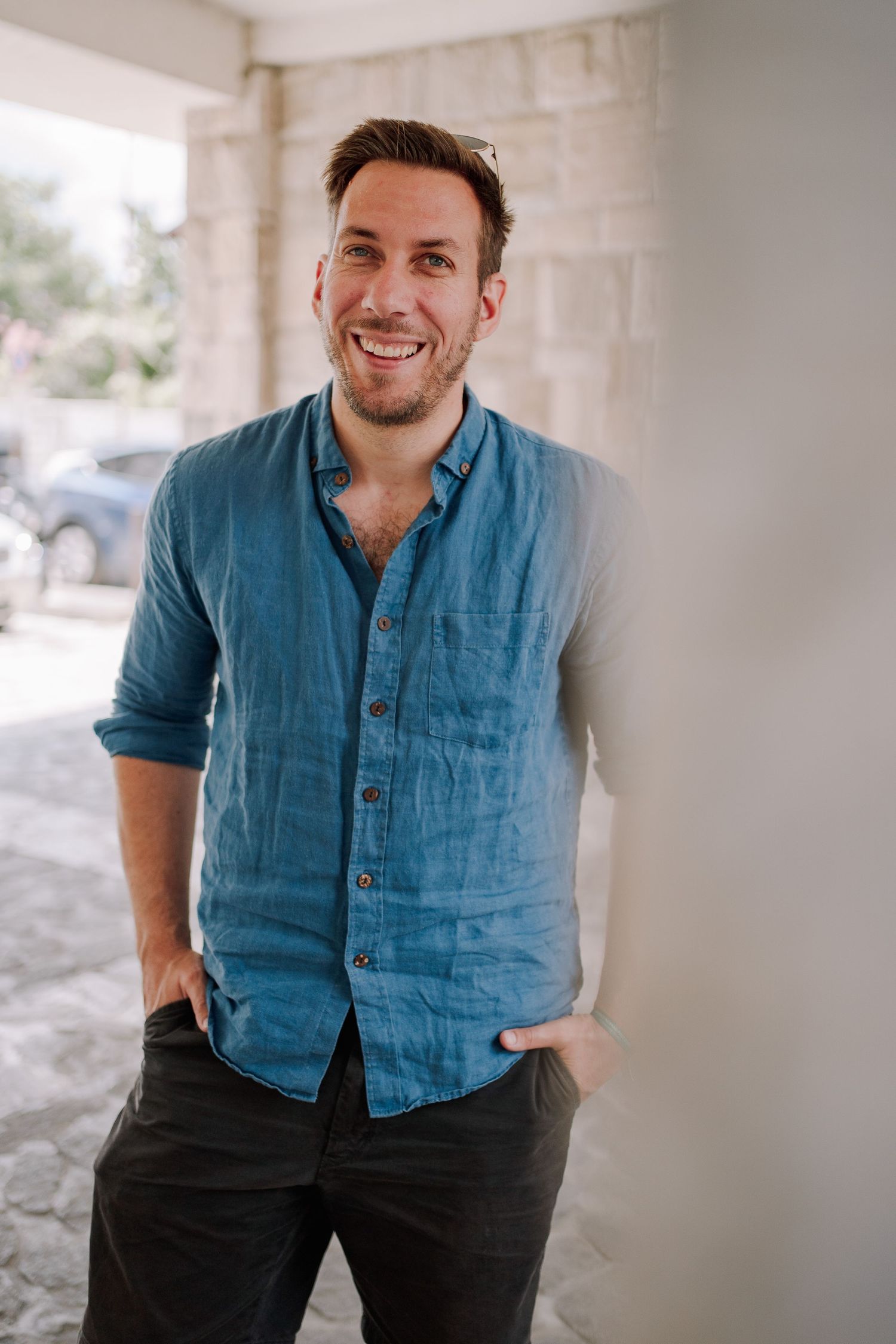 Smiling man in blue linen shirt leaning against white brick wall with hands in pockets outdoors.