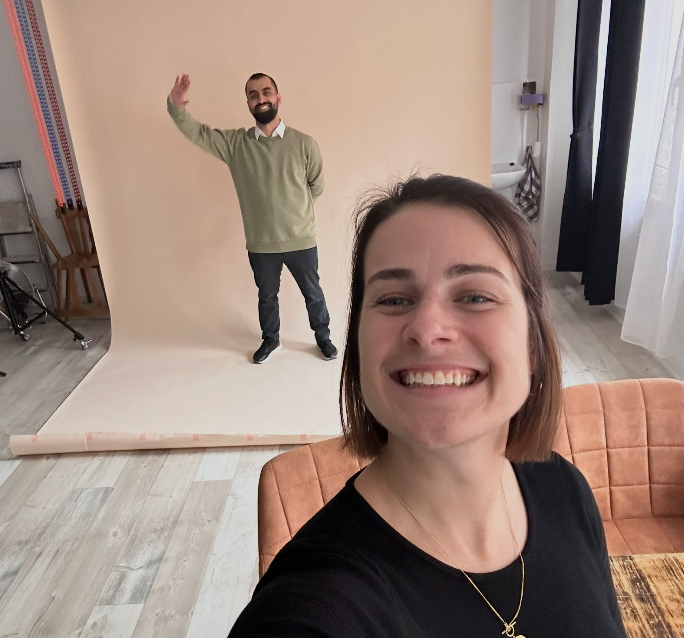A smiling woman takes a selfie in front of a man waving near a photography backdrop in a bright studio room.
