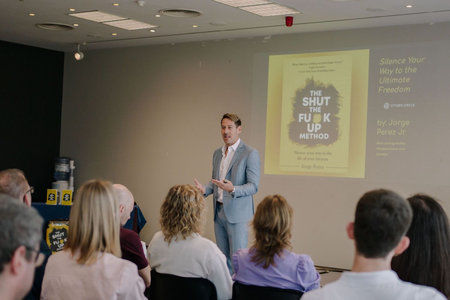 A speaker presents 'The Shut The F*ck Up Method' book at a professional seminar to an engaged audience.