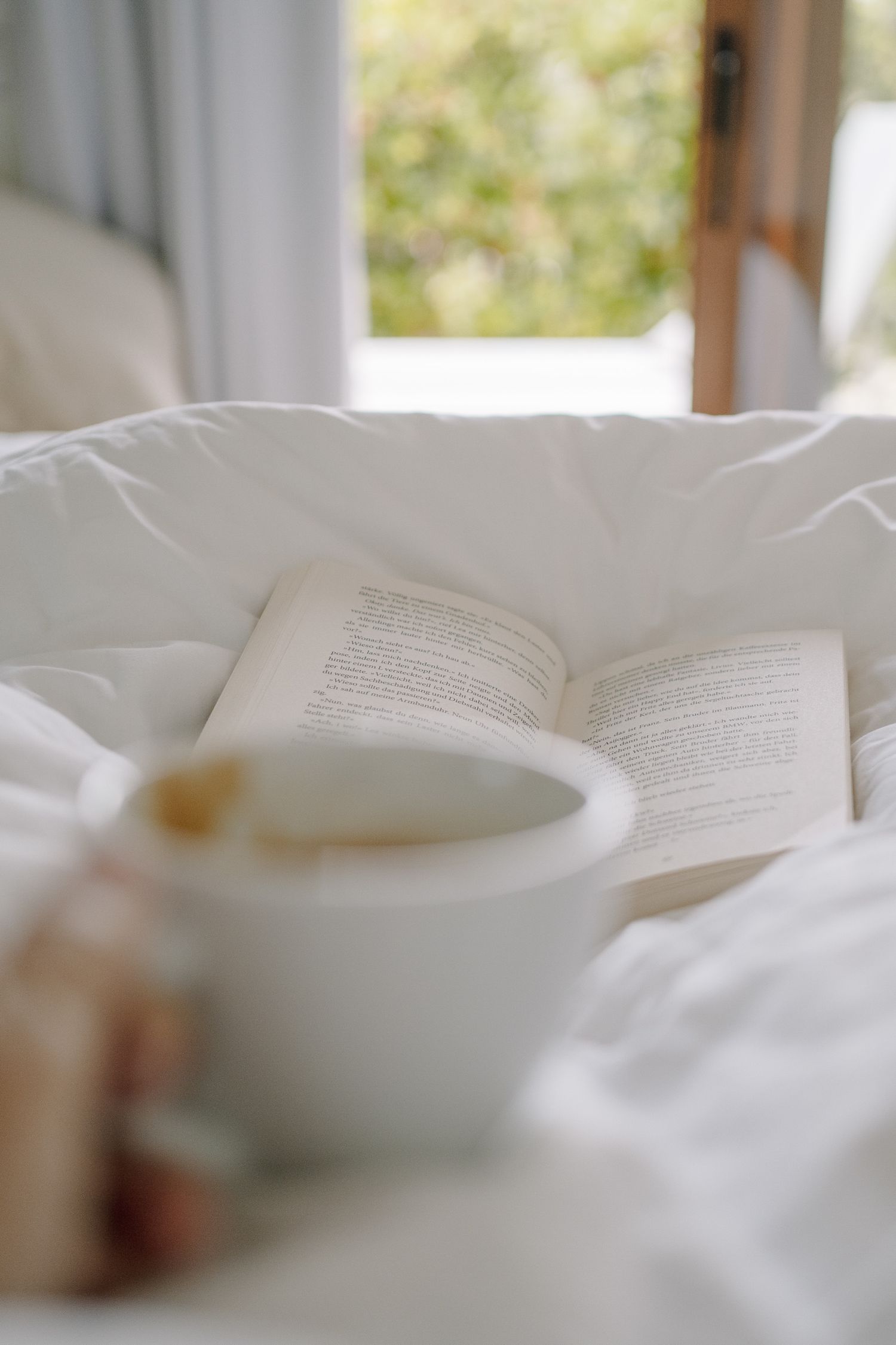 Open book resting on white bedding beside a blurred cup of coffee, with an open window and green foliage in the background.