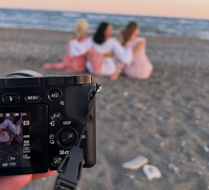 Camera screen showing three women sitting together on a sandy beach at sunset, captured in a candid photography moment.