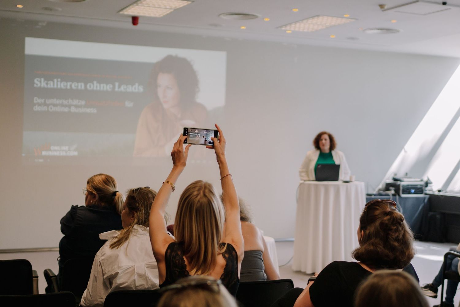 Audience members photograph a speaker presenting 'Skalieren ohne Leads' on a projector screen at a business conference.