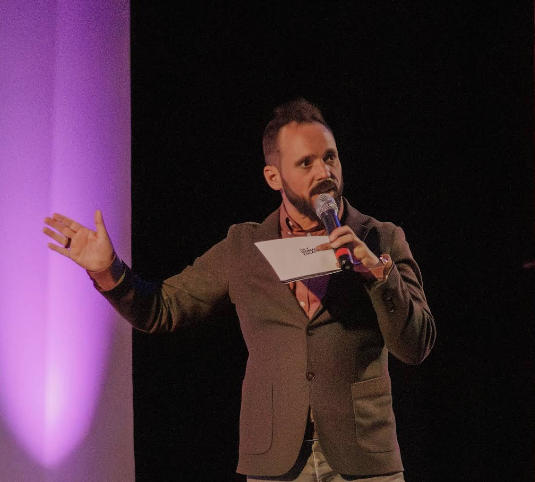 Man in gray blazer speaks into microphone on stage, gesturing with one hand under purple lighting.