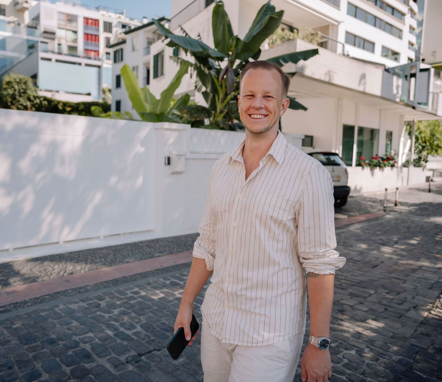 Smiling man in striped linen shirt walking outdoors on cobblestone street with tropical plants and white buildings.