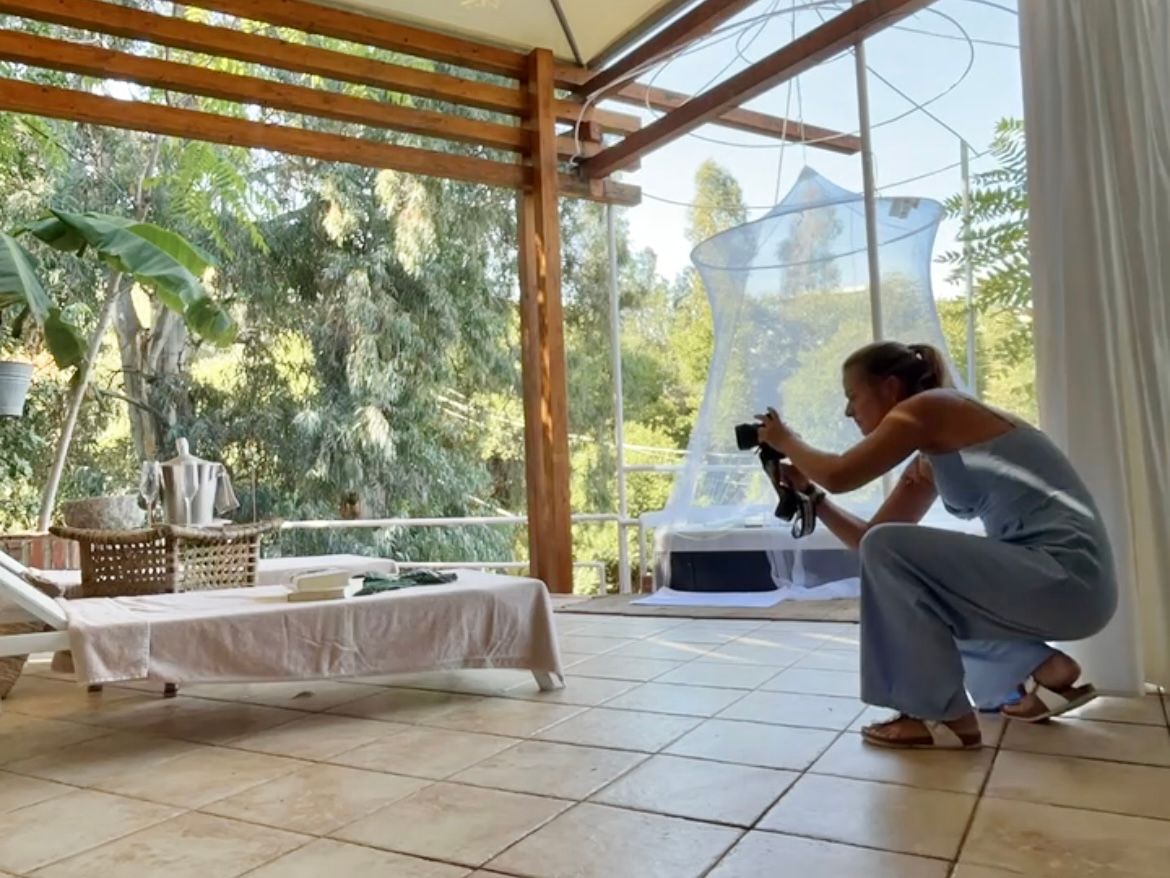Woman in a gray dress crouches on a tile floor photographing a lounge chair in a bright glass-walled room surrounded by lush green trees.