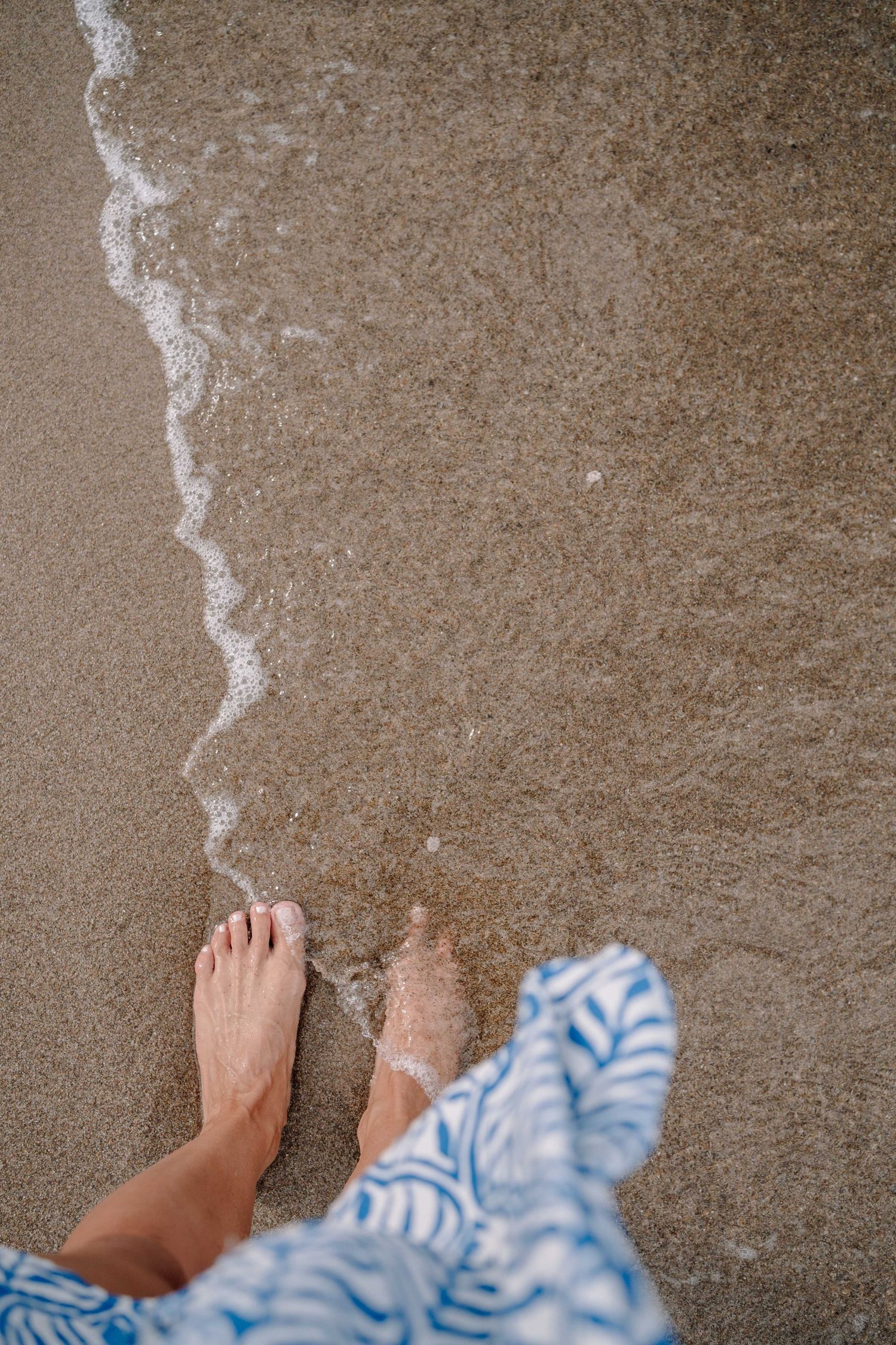 Top-down view of bare feet standing on wet sand as a gentle foamy wave washes in, with a blue and white patterned dress hem nearby.