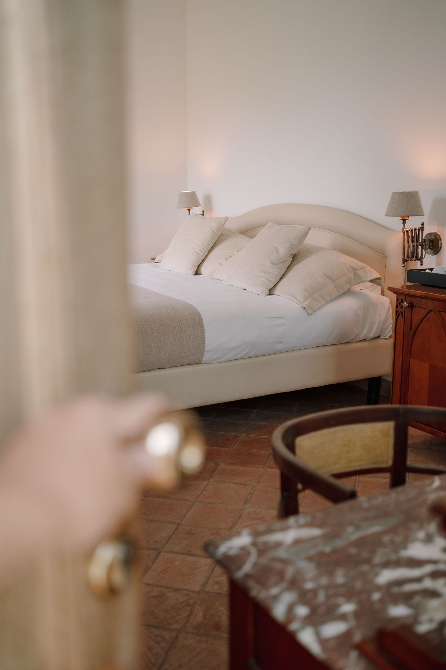 Hand opening a door to a cozy hotel bedroom with white bedding, cushions, wall sconces, terracotta tile floor and a marble-top table.