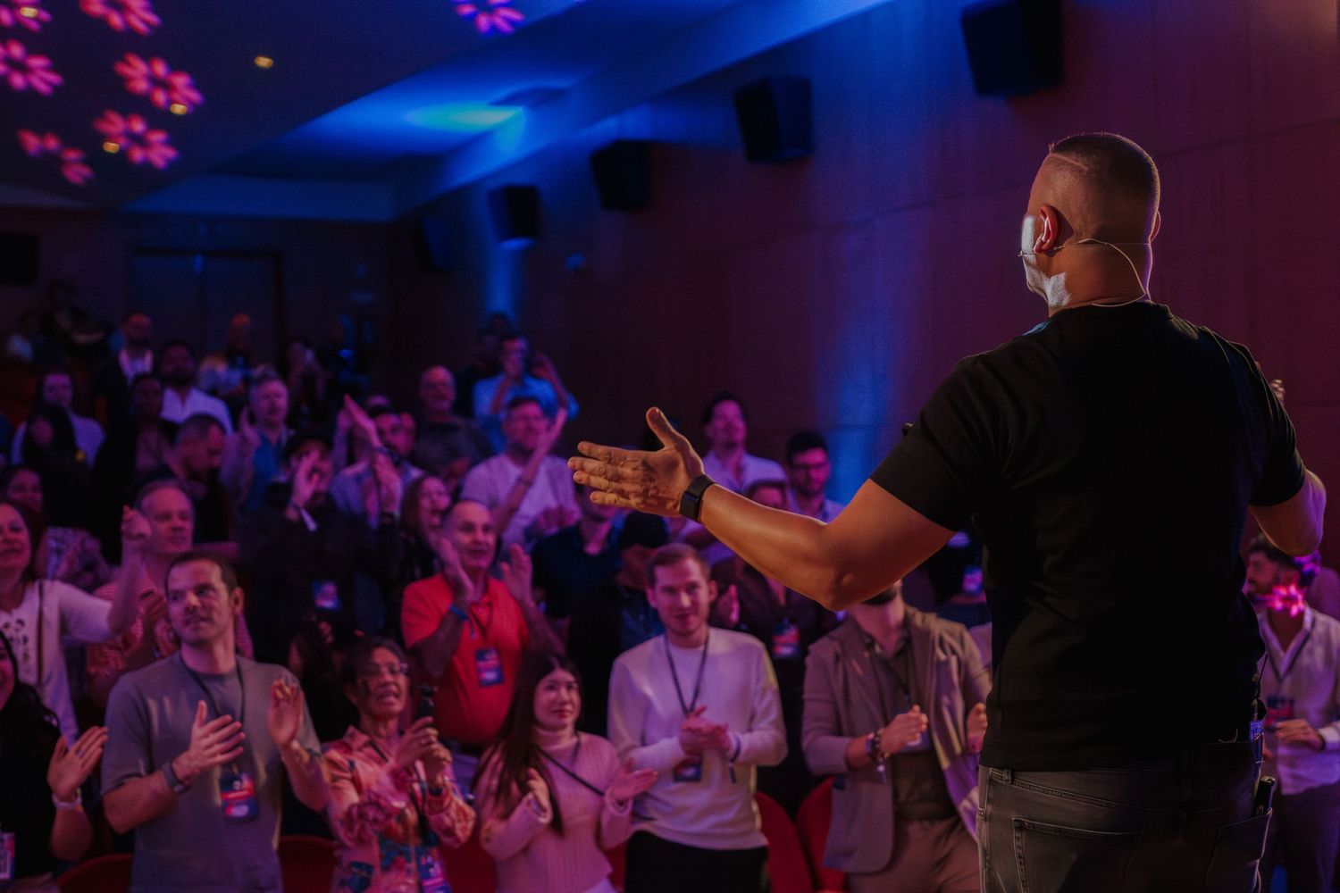 Speaker on stage facing applauding audience at a professional conference with purple lighting.